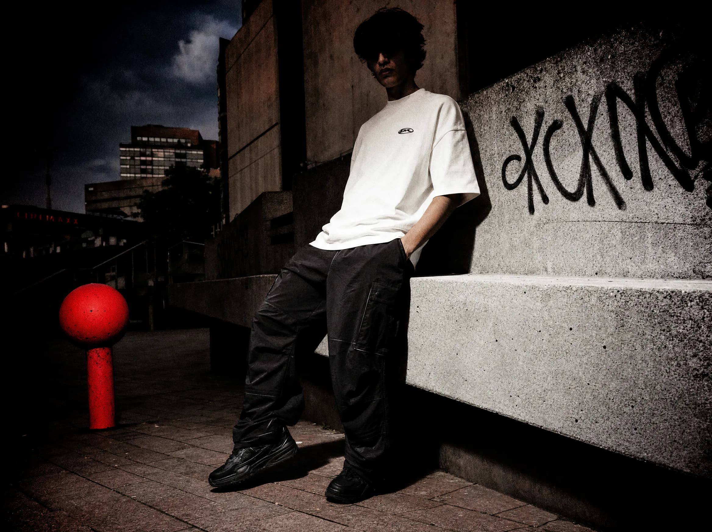 Una figura con camiseta color hueso ante un muro con grafitis en una plaza brutalista al anochecer
