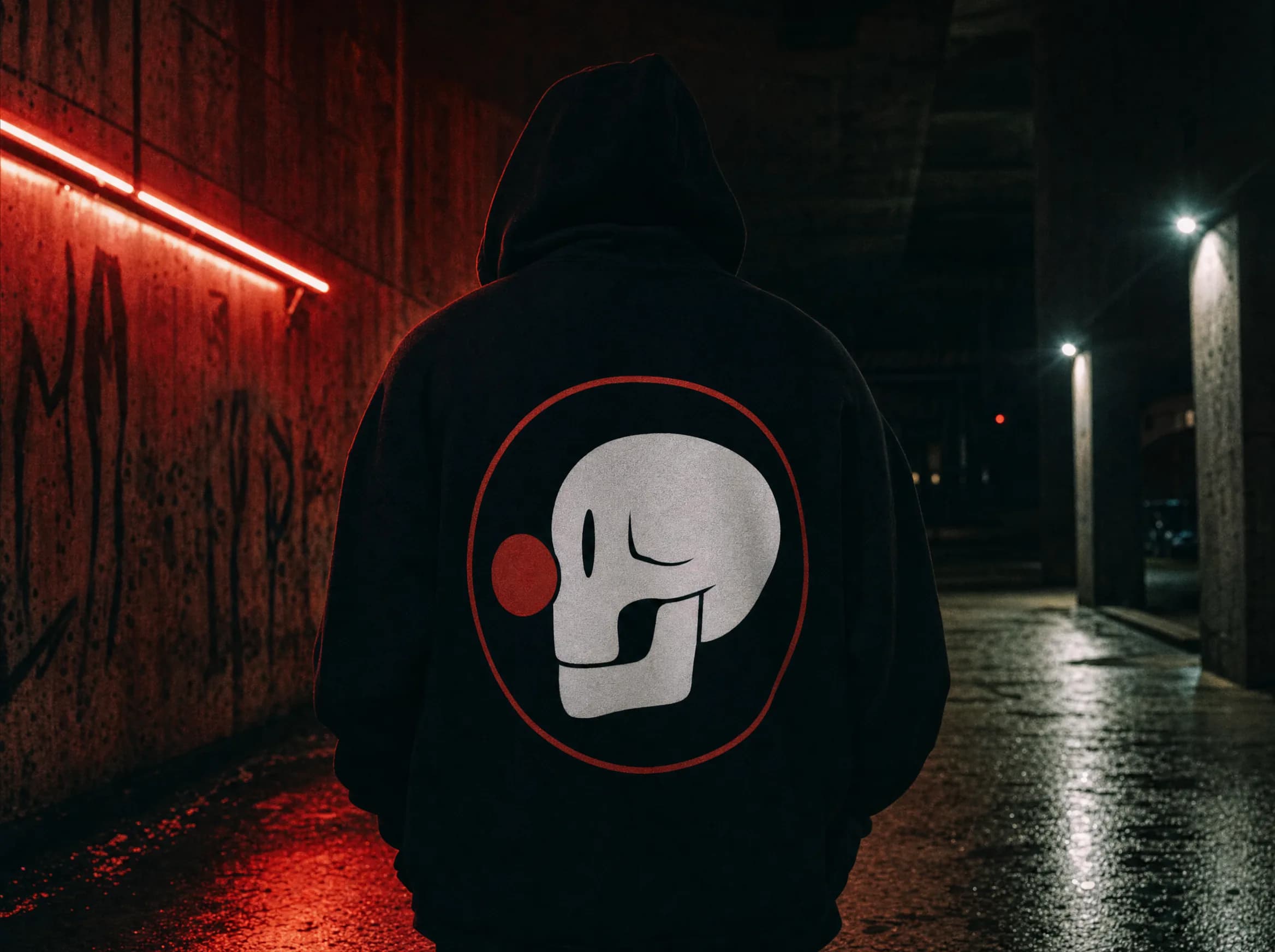 Una figura con sudadera negra oversize en un paso subterráneo de hormigón mojado bajo neón rojo